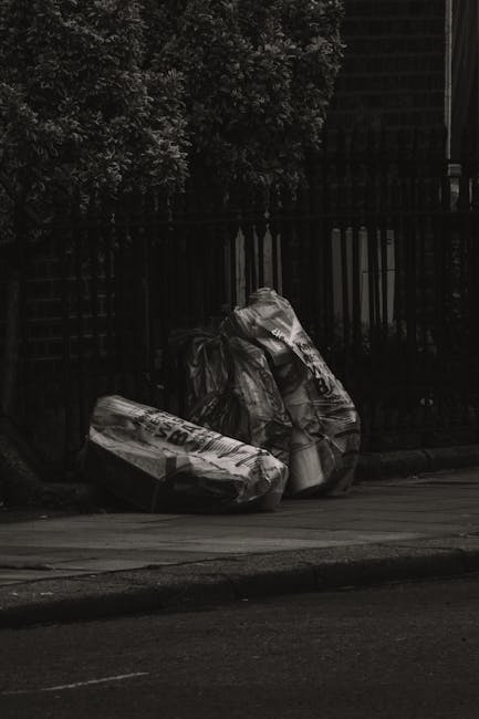 Three large black plastic rubbish bags and a partially collapsed cardboard box, all visibly crumpled and sealed, are placed on the sidewalk near a dark metal fence with vertical bars and a wooden latt