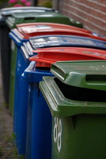 A row of large, rectangular wheelie bins positioned outdoors along a paved pathway next to a brick wall. The bins are made of heavy-duty plastic with textured surfaces on the lids and sides. The front
