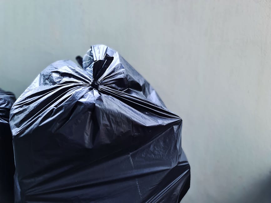 A row of large, rectangular wheelie bins positioned outdoors along a paved pathway next to a brick wall. The bins are made of heavy-duty plastic with textured surfaces on the lids and sides. The front